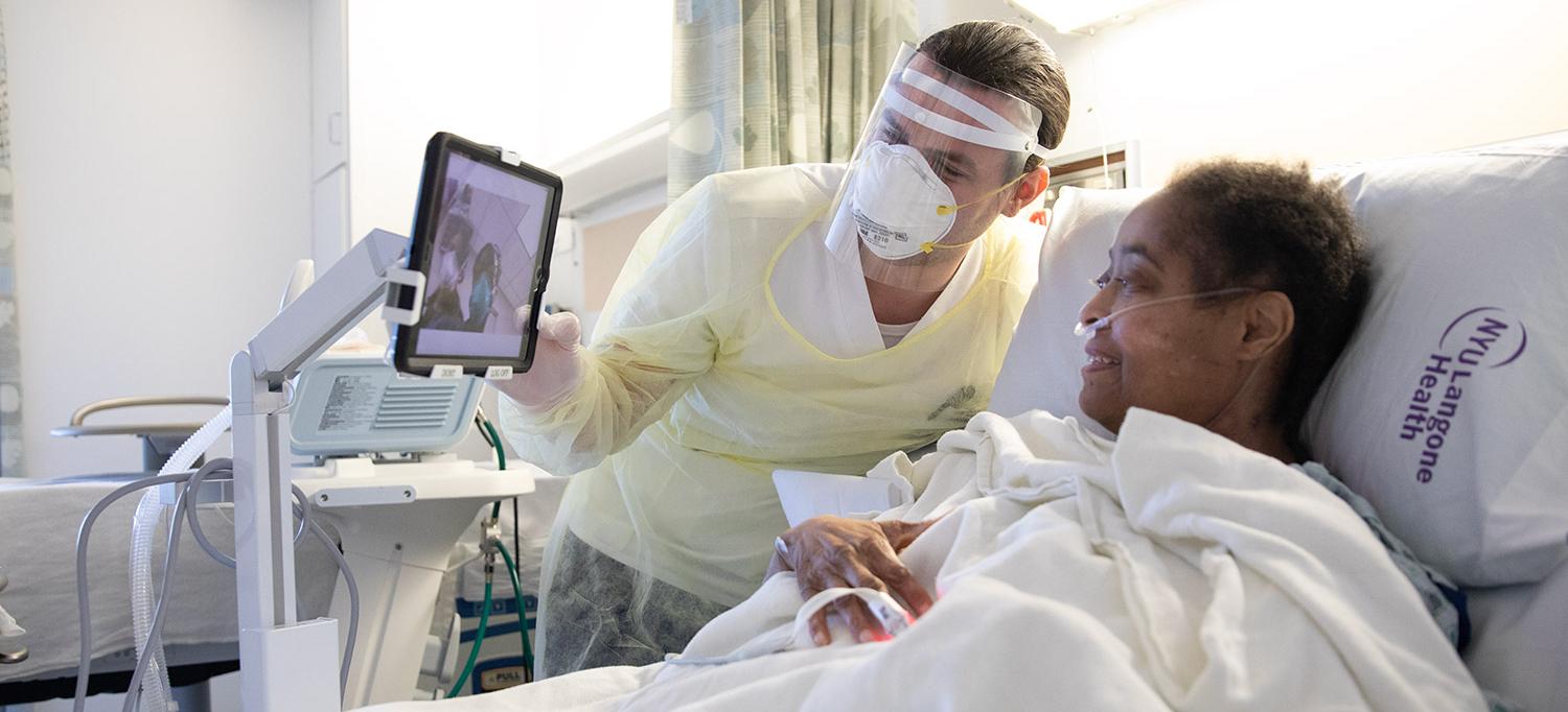 Healthcare Provider Wearing Personal Protective Equipment Helps Patient in Hospital Bed Speak with People on Tablet