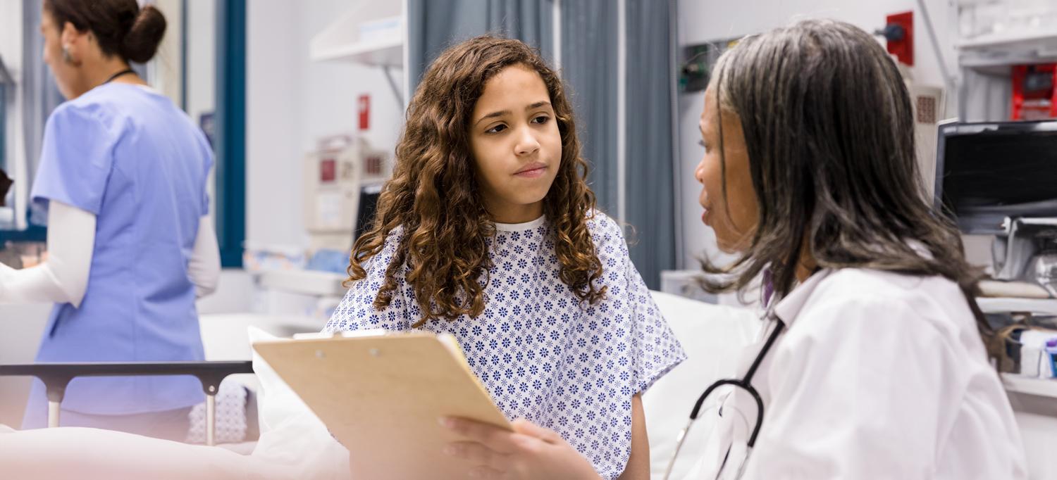 A doctor holding a clipboard speaks to a young patient, standing in front of a hospital bed.