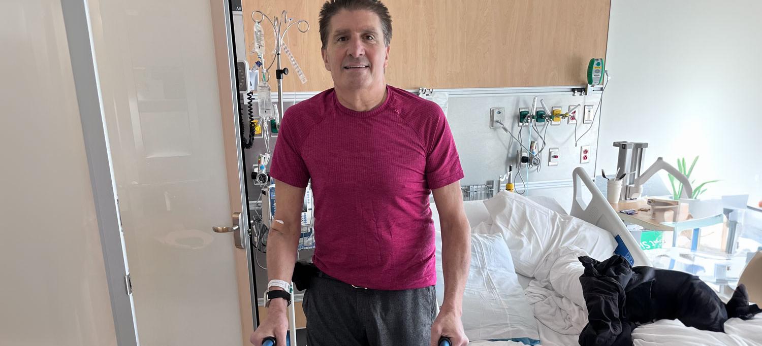 Using a walker, Richard Monti stands smiling in his hospital room.