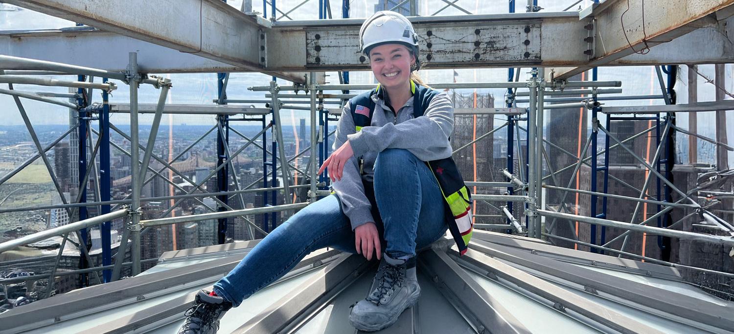 Wearing a hardhat and smiling, Oberle sits on a section of a building under construction.